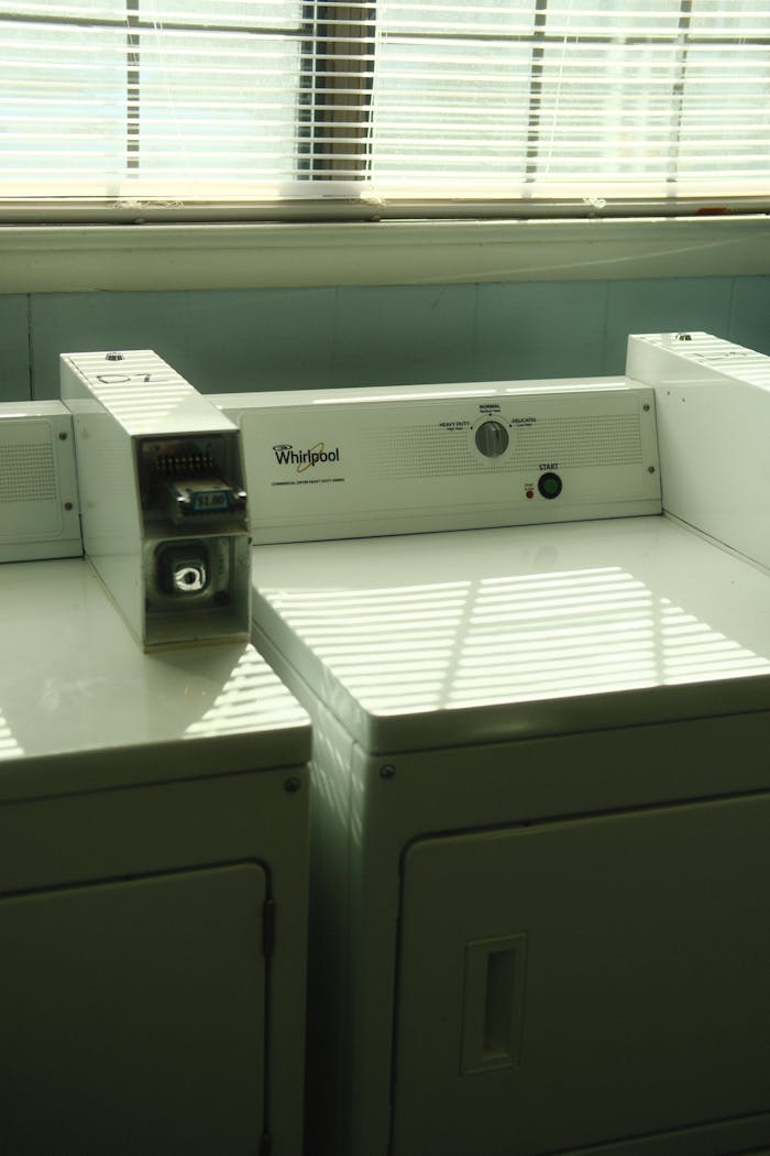 Two commercial laundry machines in a sunlit room with blinds casting shadows on surfaces.